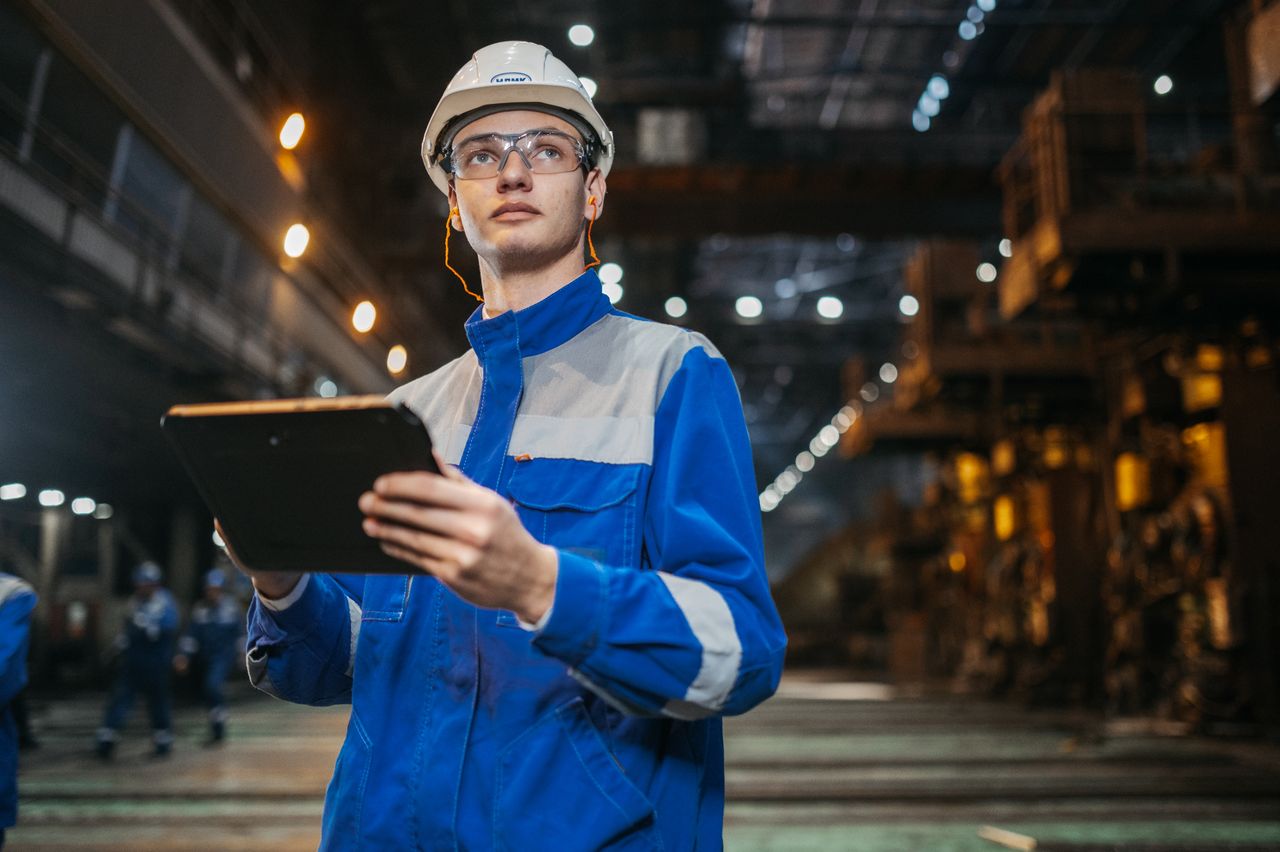Engineer in safety gear uses a tablet in a large industrial factory setting, focused and diligent.