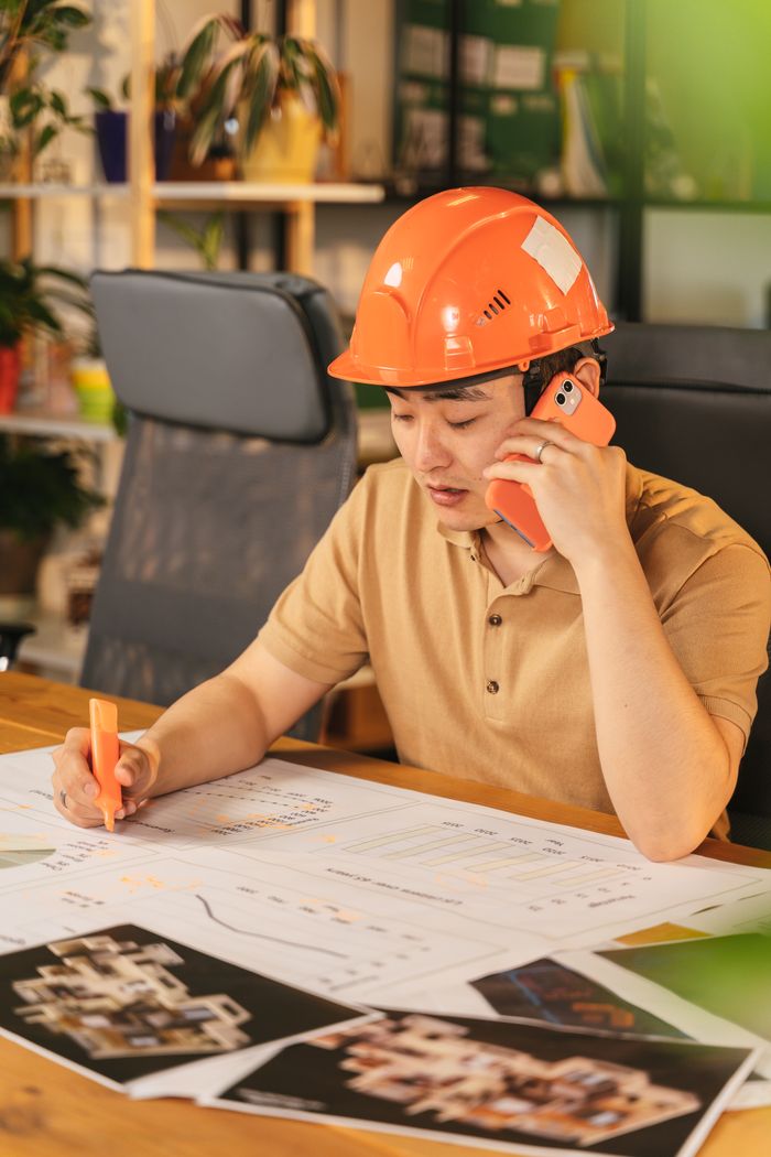 creative-03 Asian male architect reviewing blueprints and talking on phone indoors, wearing a hard hat.